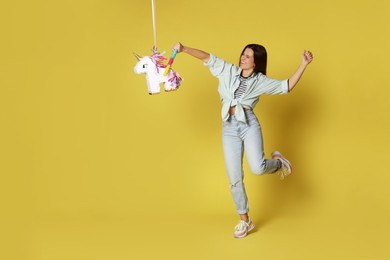 Photo of Happy woman hitting unicorn shaped pinata with stick on yellow background