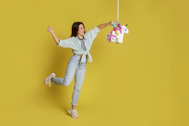 Photo of Happy woman hitting unicorn shaped pinata with stick on yellow background