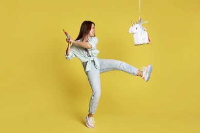 Photo of Happy woman hitting unicorn shaped pinata with stick on yellow background