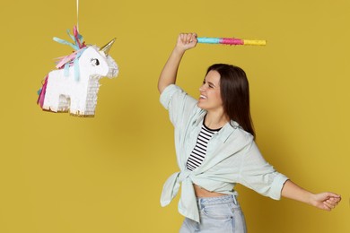 Happy woman hitting unicorn shaped pinata with stick on yellow background