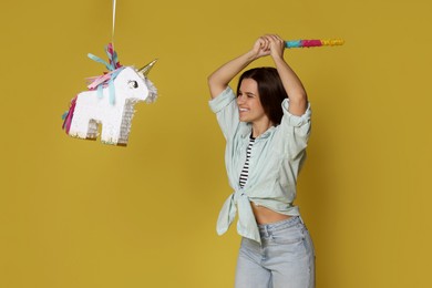 Photo of Happy woman hitting unicorn shaped pinata with stick on yellow background