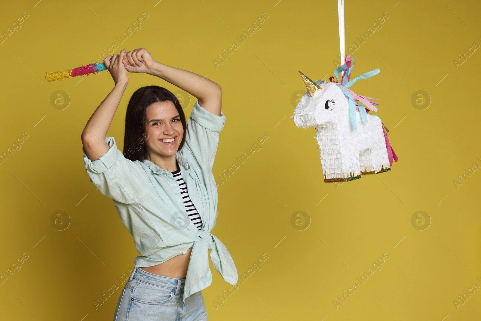 Photo of Happy woman hitting unicorn shaped pinata with stick on yellow background