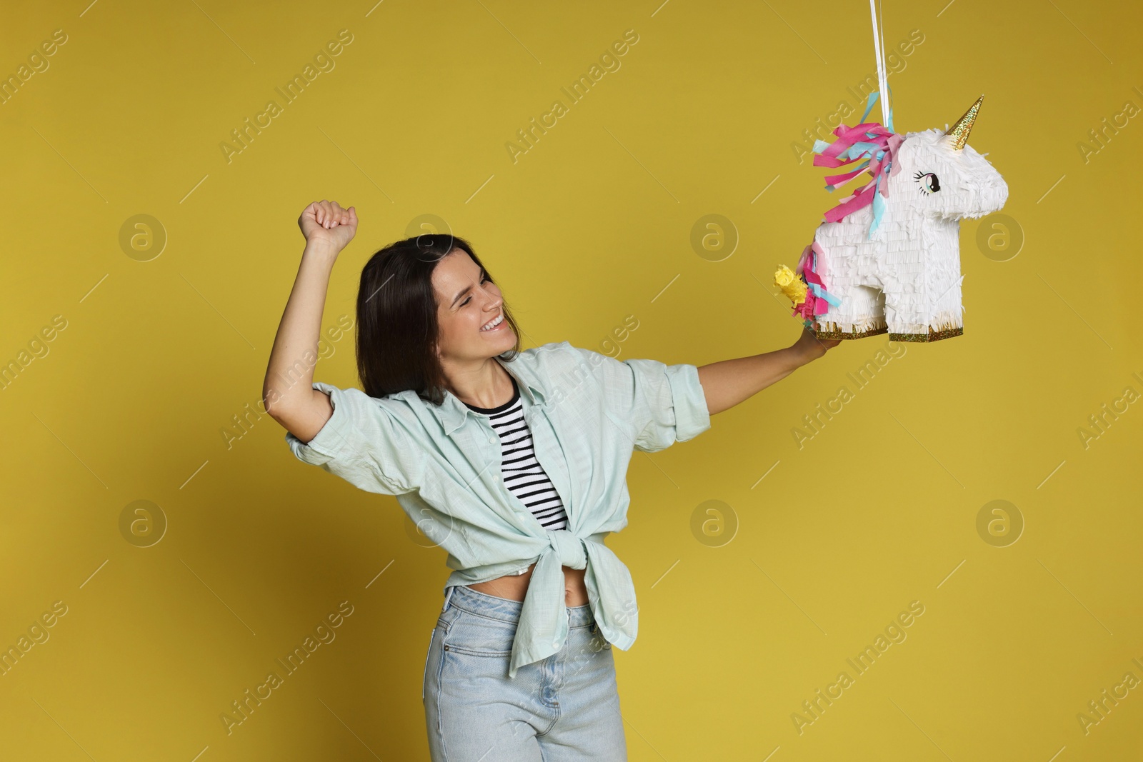 Photo of Happy woman hitting unicorn shaped pinata with stick on yellow background