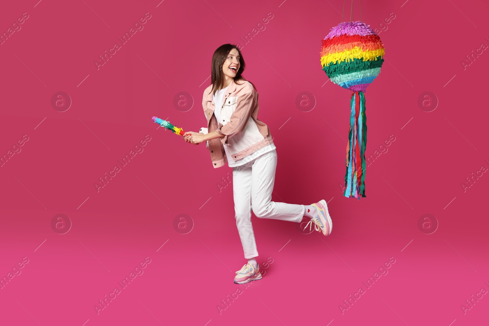 Photo of Happy woman breaking colorful pinata with stick on pink background