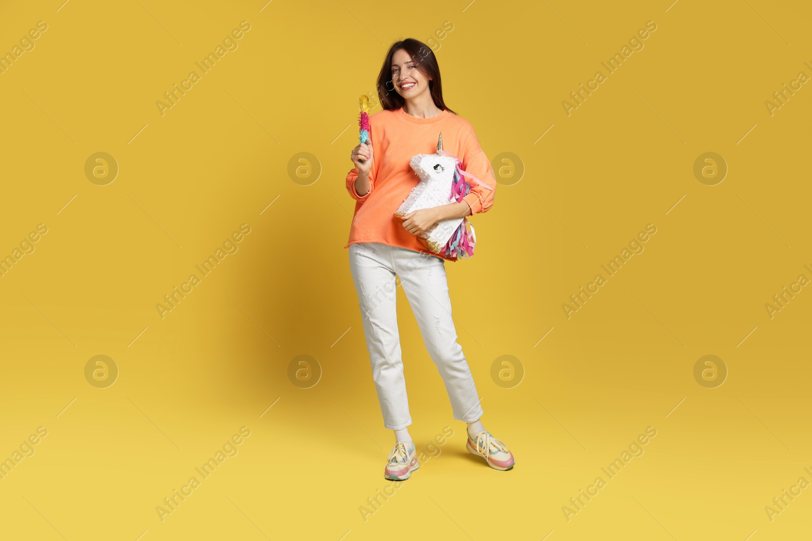 Photo of Happy woman with unicorn shaped pinata and stick on yellow background