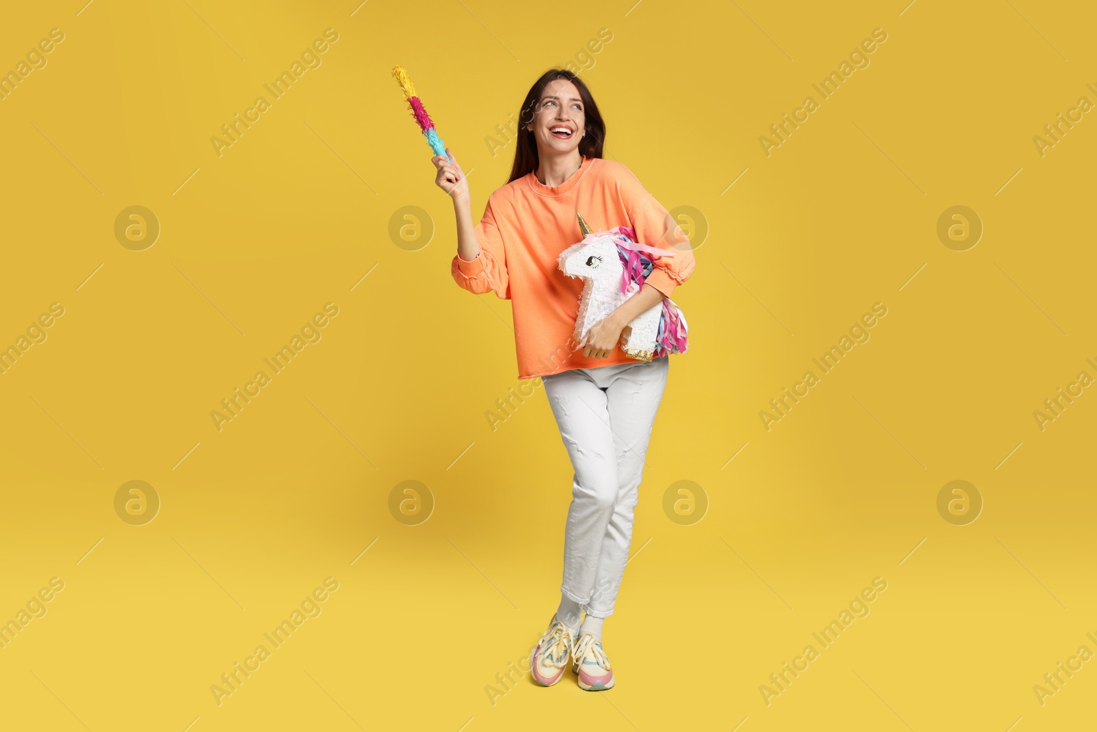 Photo of Happy woman with unicorn shaped pinata and stick on yellow background