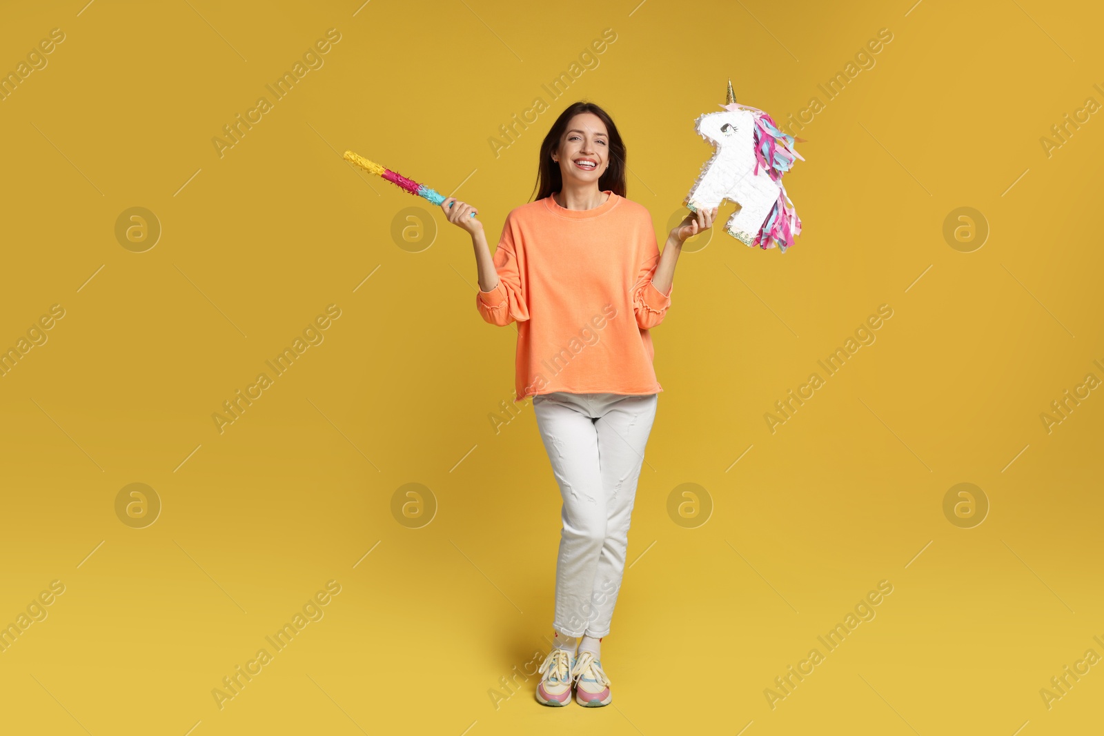 Photo of Happy woman with unicorn shaped pinata and stick on yellow background