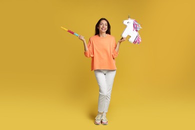 Happy woman with unicorn shaped pinata and stick on yellow background