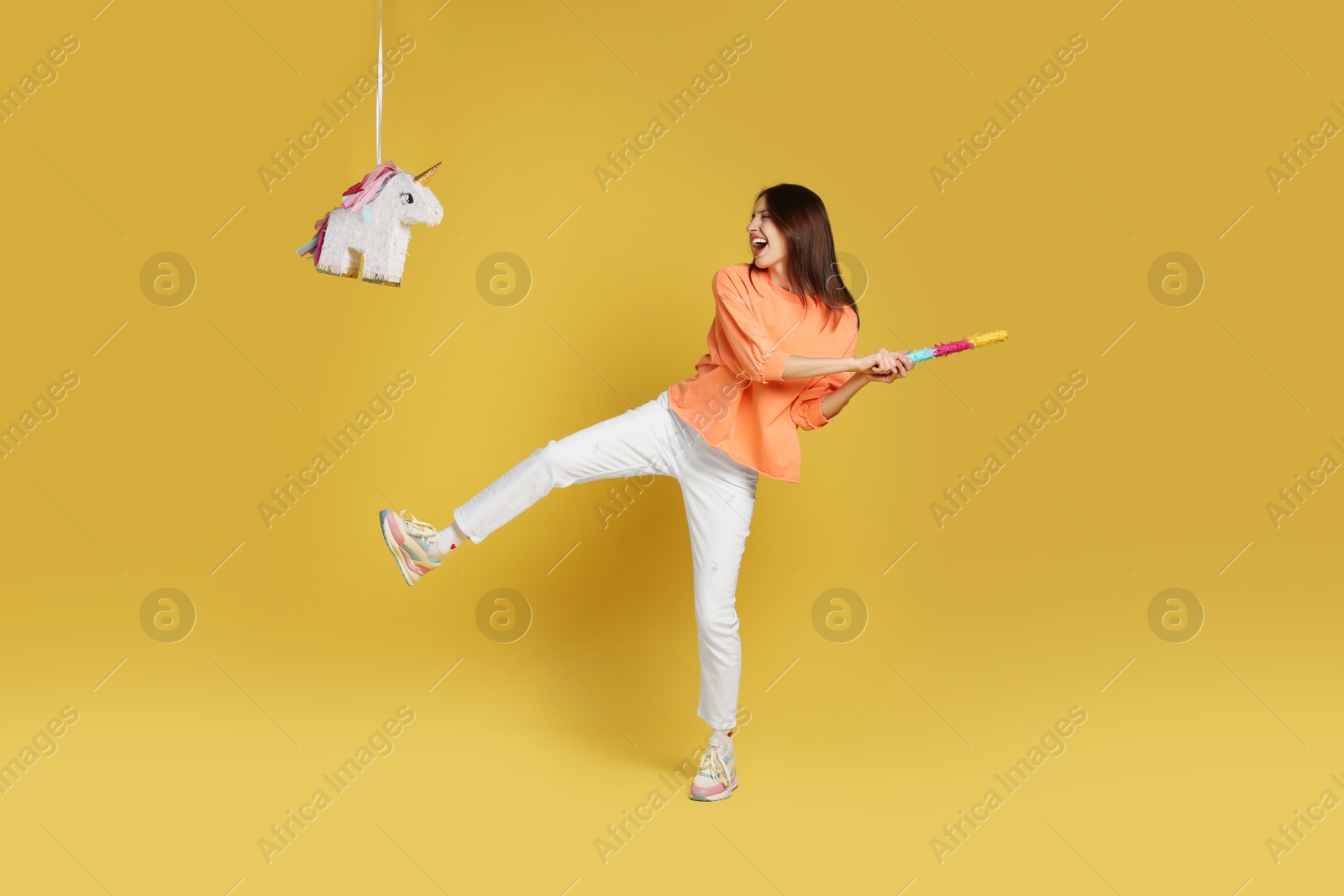 Photo of Happy woman breaking unicorn shaped pinata with stick on yellow background