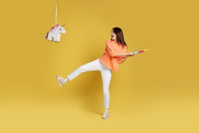 Photo of Happy woman breaking unicorn shaped pinata with stick on yellow background