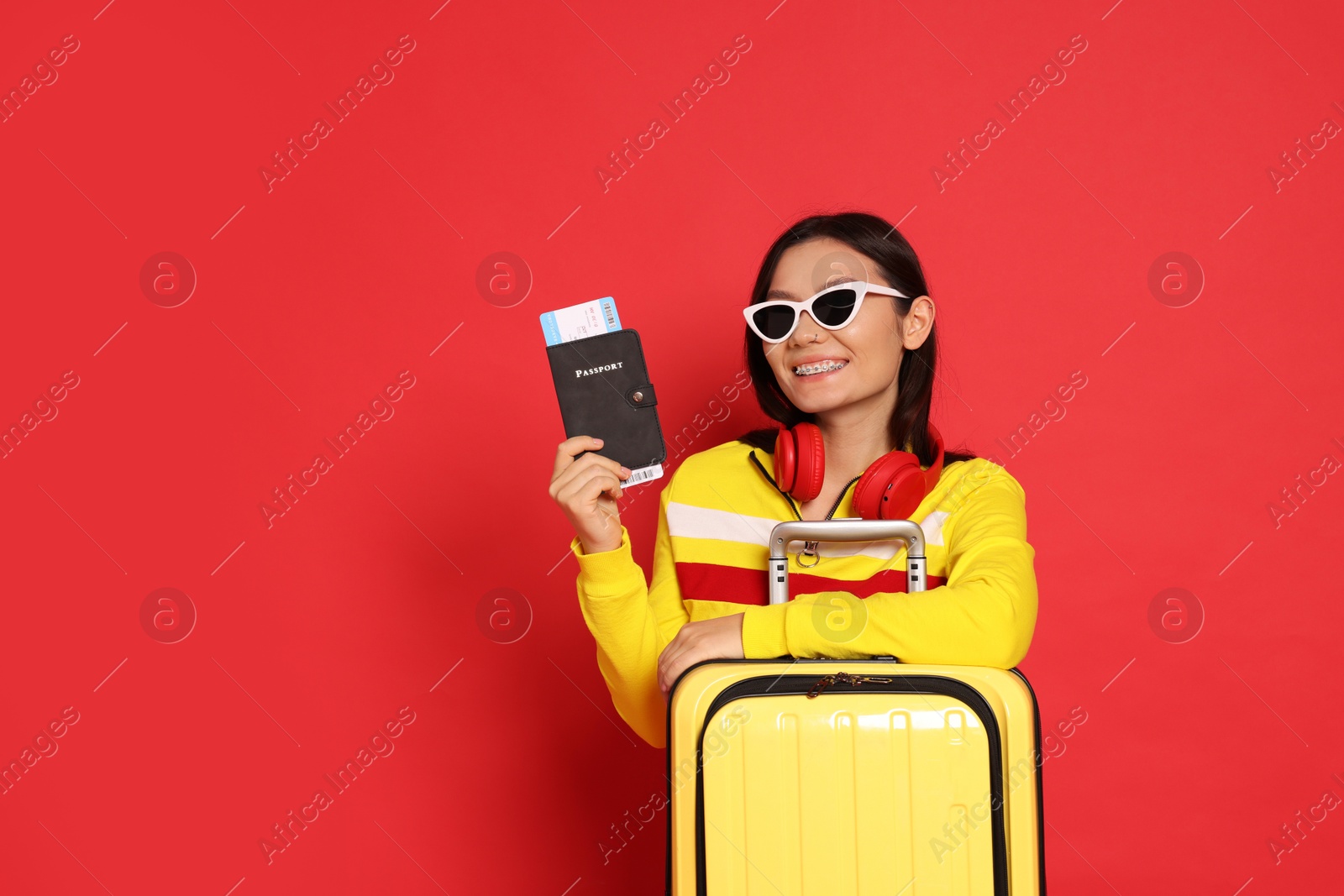 Photo of Happy traveller with headphones, passport and suitcase on red background. Space for text