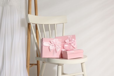 Photo of Gift boxes for wedding day on chair and wooden rack with bridal dress near white wall, space for text