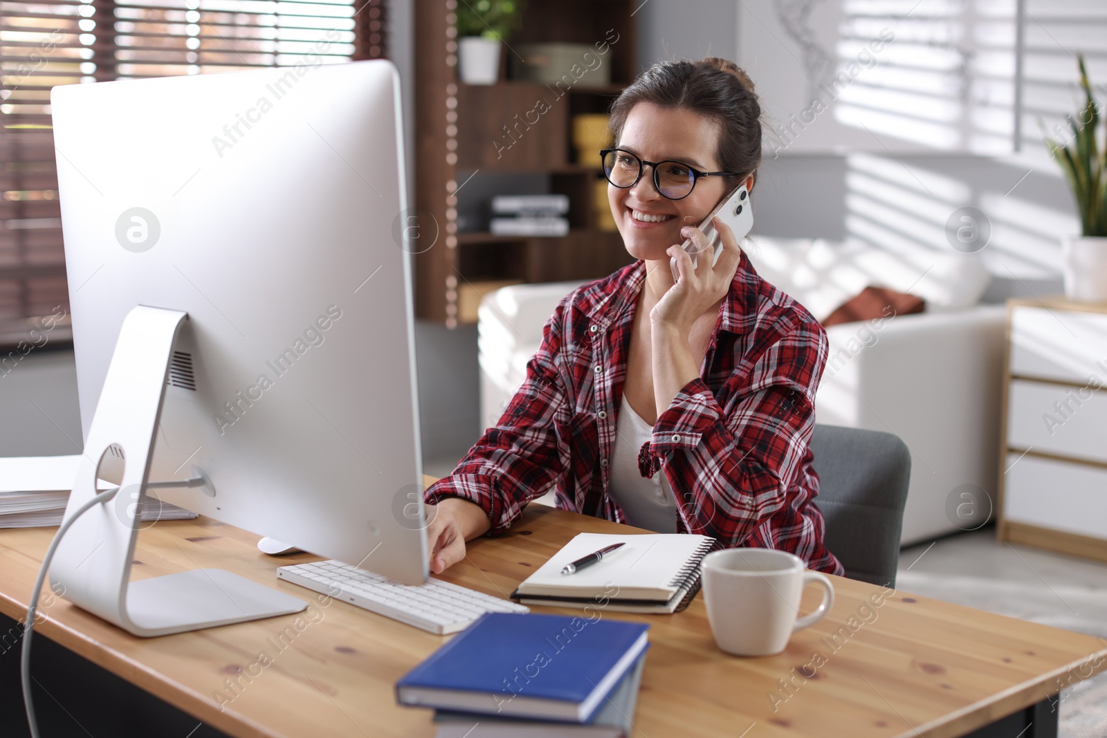 Photo of Happy woman talking by smartphone at desk with computer indoors. Home office