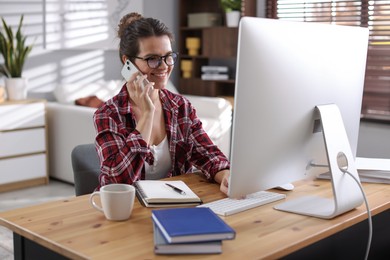 Happy woman talking by smartphone at desk with computer indoors. Home office