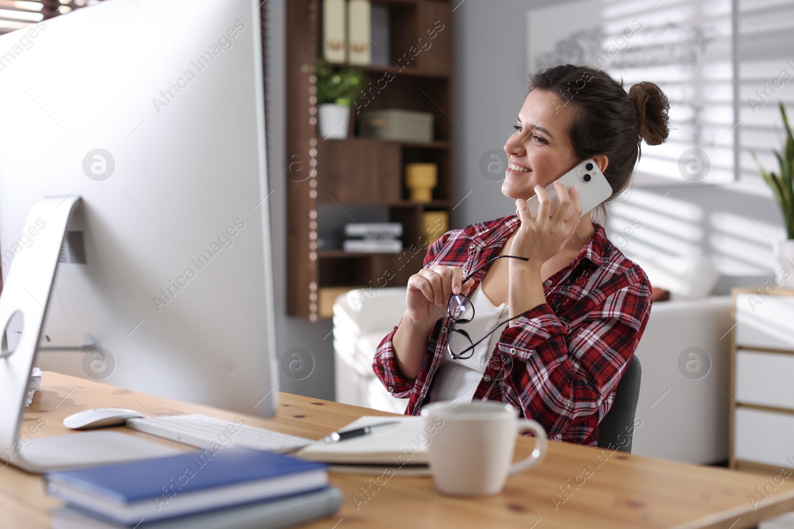 Photo of Happy woman talking by smartphone at desk with computer indoors. Home office