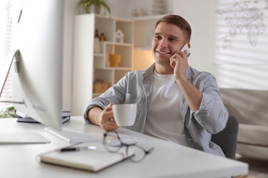 Photo of Happy man talking by smartphone at desk with computer indoors. Home office