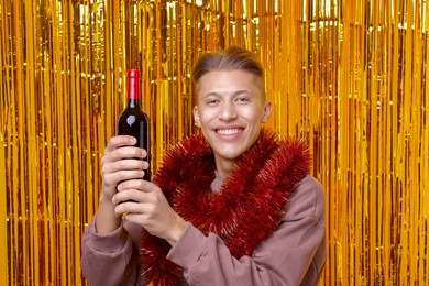 Photo of Happy young man with bright tinsel and bottle of wine against golden foil curtain