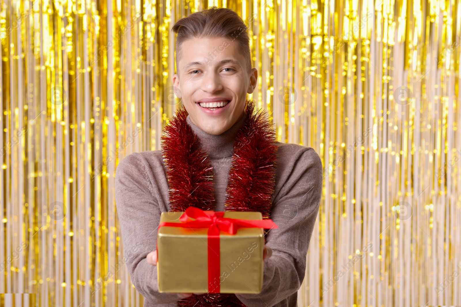 Photo of Happy young man with bright tinsel and gift box against foil curtain