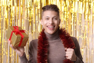 Photo of Happy young man with bright tinsel and gift box against foil curtain