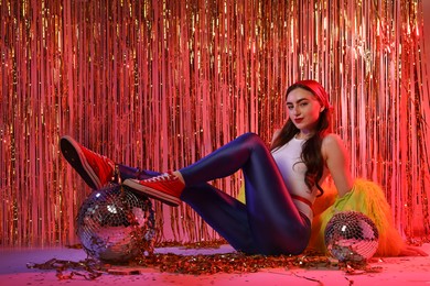 Photo of Beautiful young woman wearing retro outfit with tinsel and disco balls against golden foil curtain in neon lights