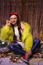 Photo of Beautiful young woman in retro outfit with shiny tinsel and disco balls indoors