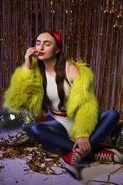 Beautiful young woman in retro outfit with shiny tinsel and disco balls indoors