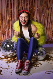 Photo of Emotional young woman in retro outfit with shiny tinsel and disco balls indoors