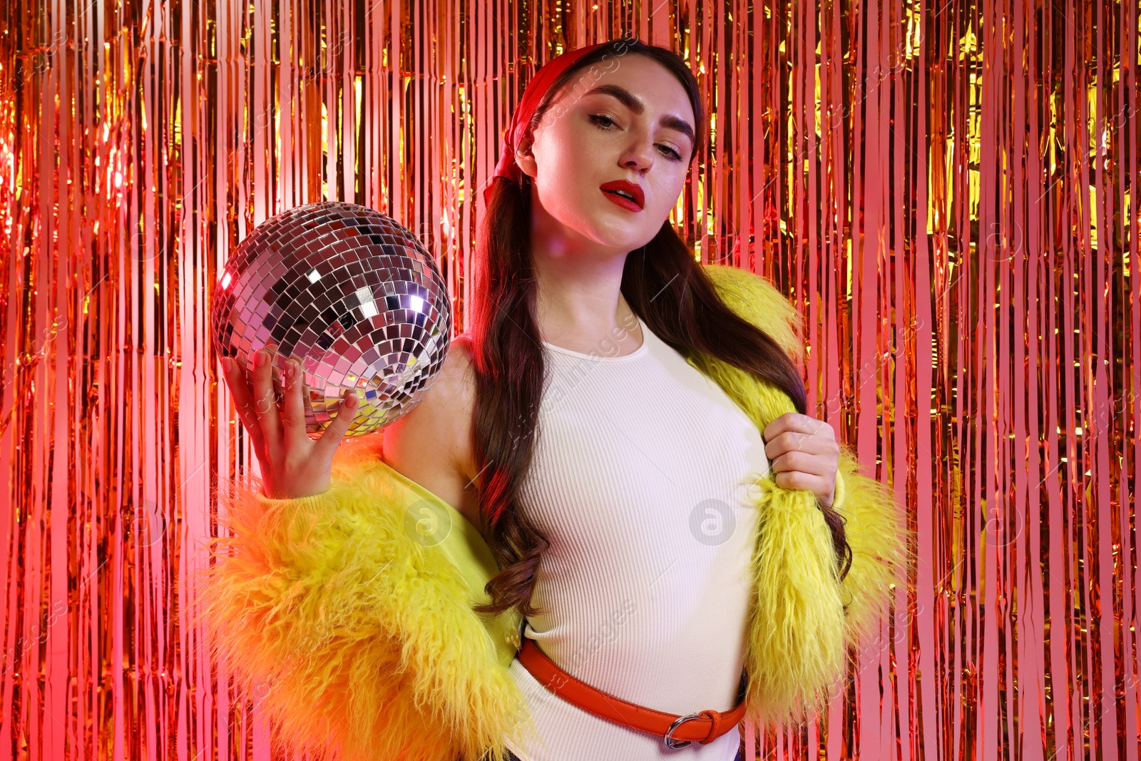 Photo of Beautiful young woman in faux fur coat with disco ball against golden foil curtain