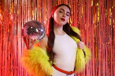 Photo of Beautiful young woman in faux fur coat with disco ball against golden foil curtain