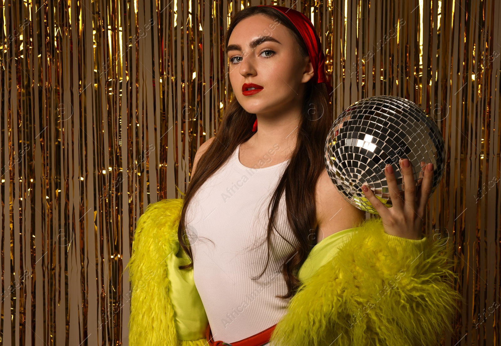 Photo of Beautiful young woman in faux fur coat with disco ball against golden foil curtain