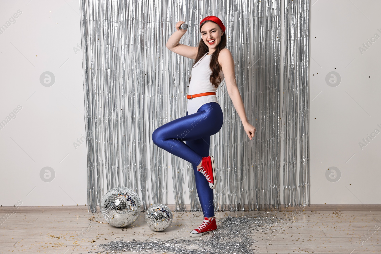 Photo of Beautiful young woman in retro outfit with microphone near silver foil curtain indoors