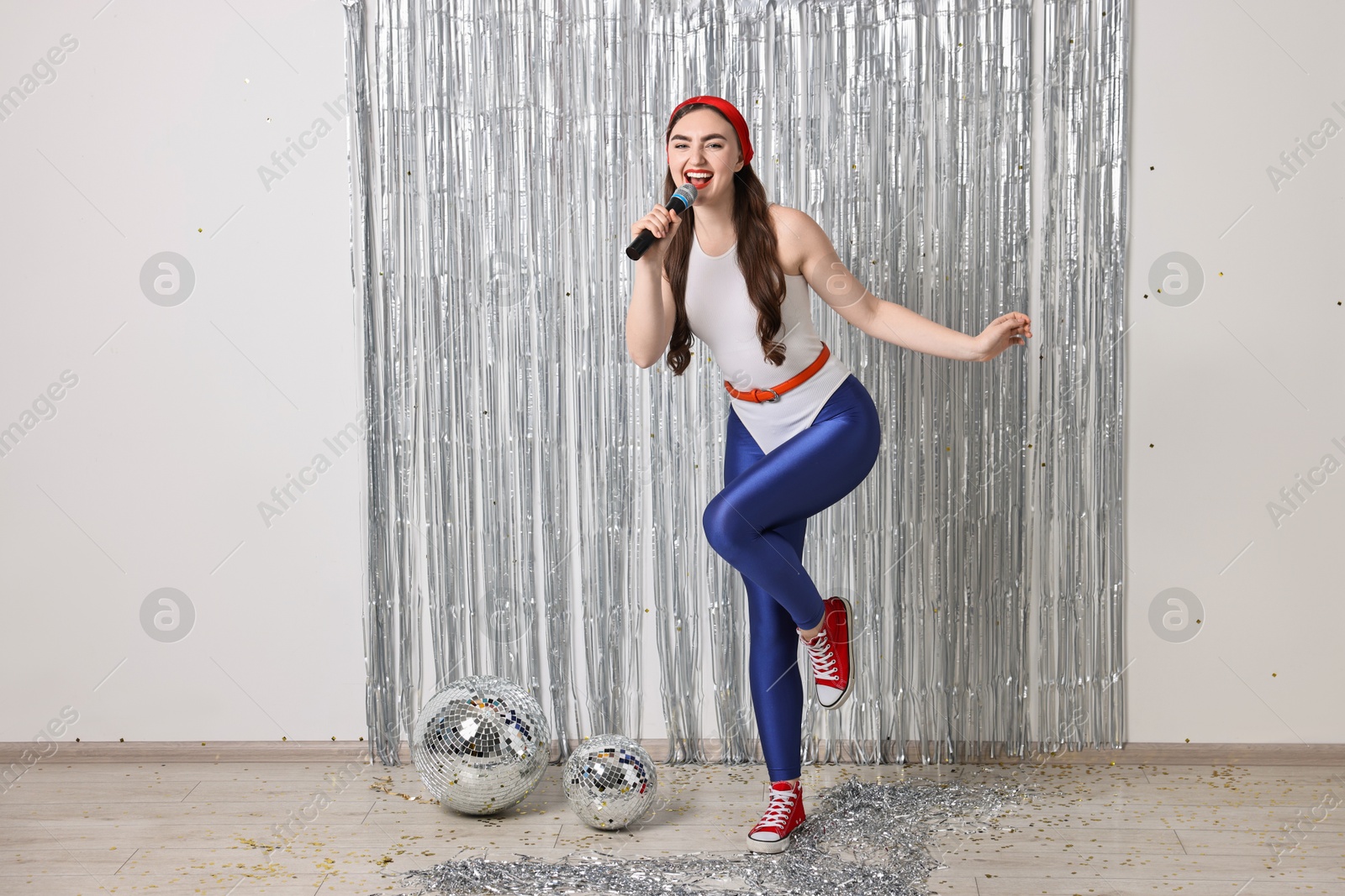 Photo of Beautiful young woman in retro outfit with microphone near silver foil curtain indoors