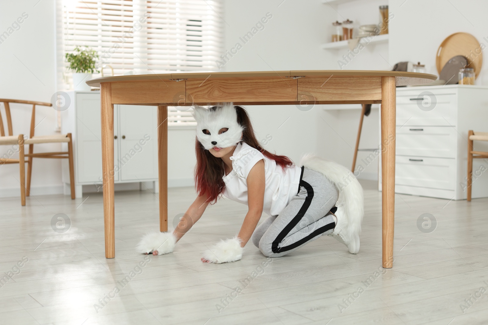 Photo of Quadrobics. Girl wearing cat mask, tail and gloves under table at home