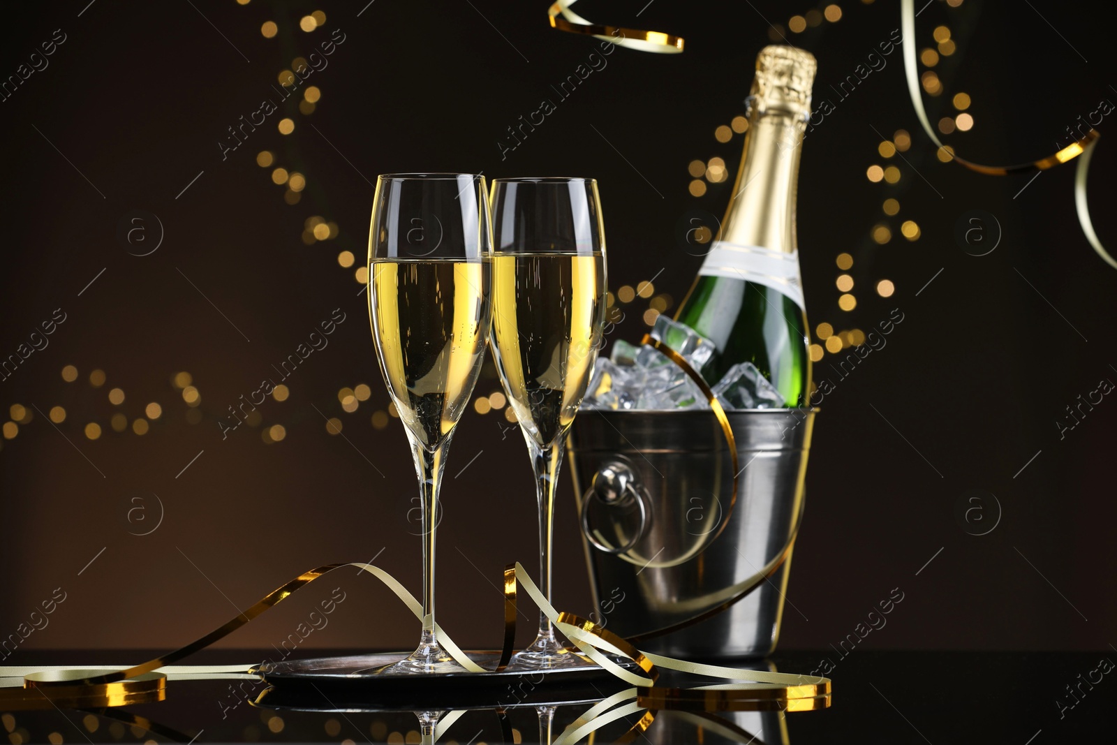 Photo of Champagne in glasses and ice bucket with bottle on mirror surface against brown background with blurred lights