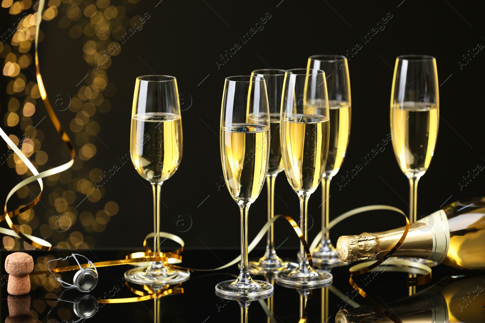 Photo of Champagne in glasses, bottle and cork on mirror surface against black background with blurred lights