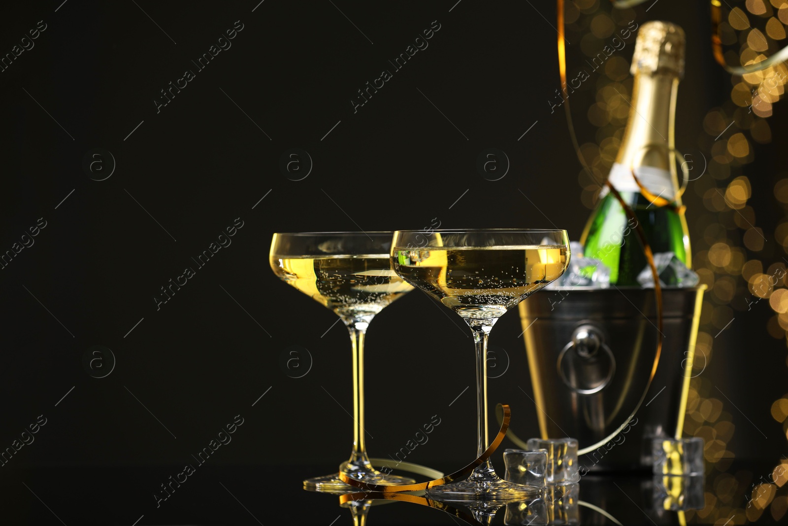 Photo of Champagne in glasses and ice bucket with bottle on mirror surface against black background with blurred lights, space for text