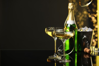 Photo of Champagne in glasses and ice bucket with bottle on mirror surface against black background with blurred lights, space for text