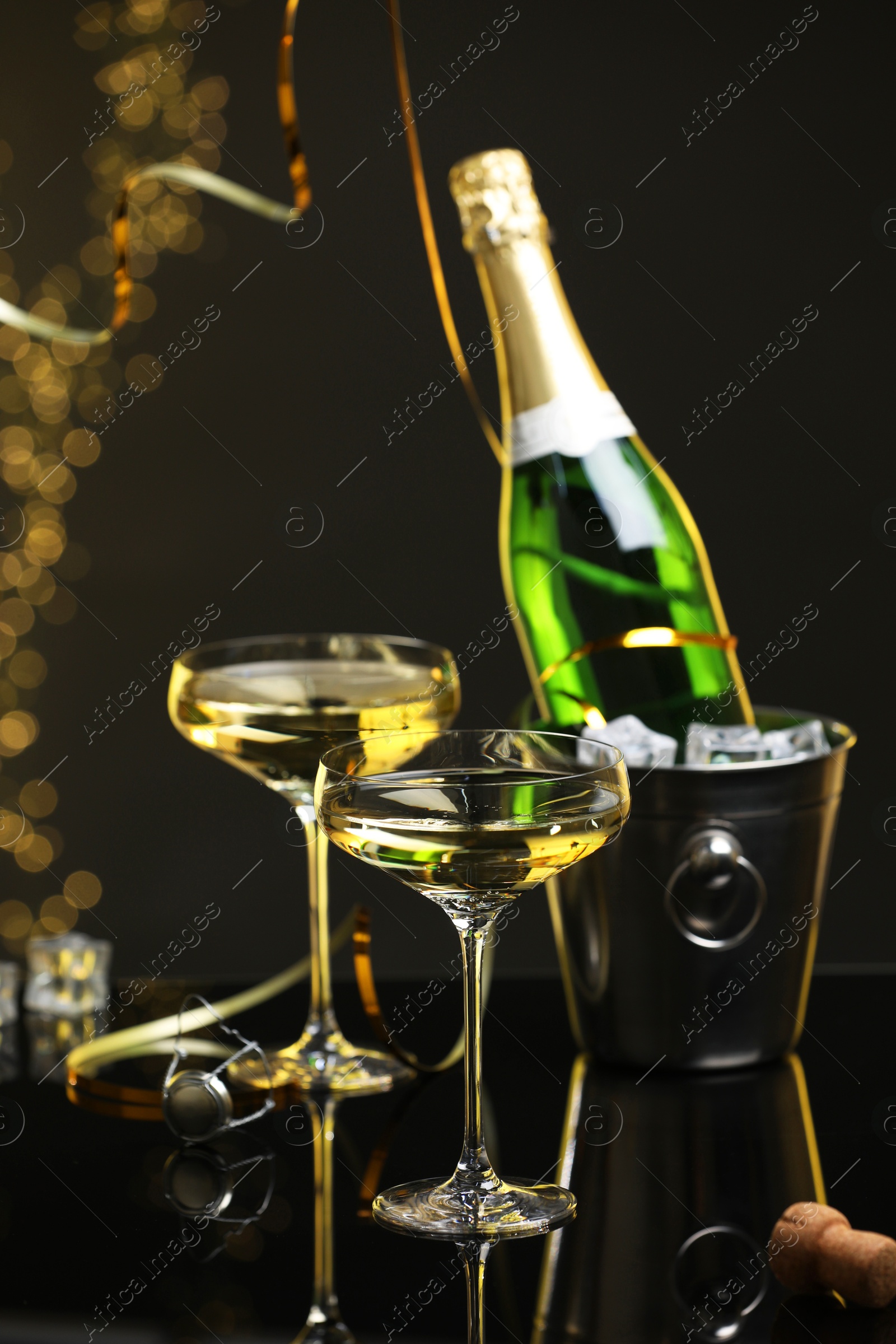 Photo of Champagne in glasses and ice bucket with bottle on mirror surface against black background with blurred lights