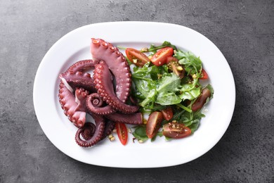 Photo of Plate with tasty boiled octopus tentacles and salad on grey textured table, top view