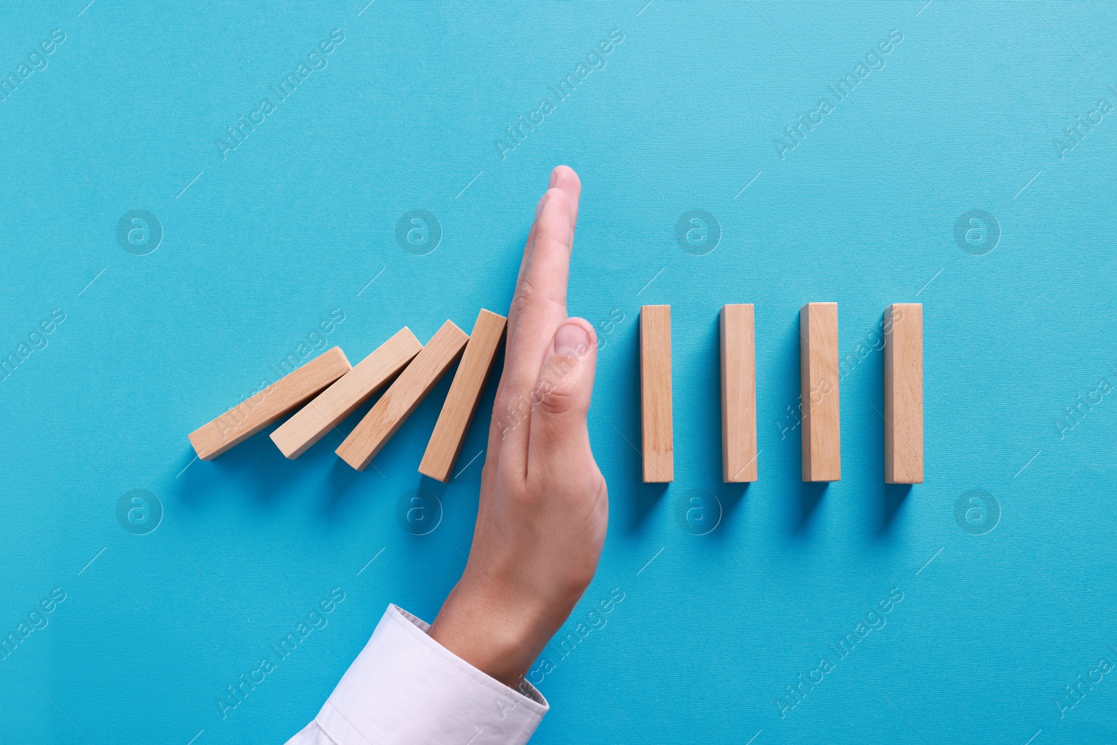 Photo of Man stopping wooden blocks from falling on light blue background, top view. Domino effect