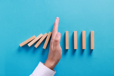 Photo of Man stopping wooden blocks from falling on light blue background, top view. Domino effect