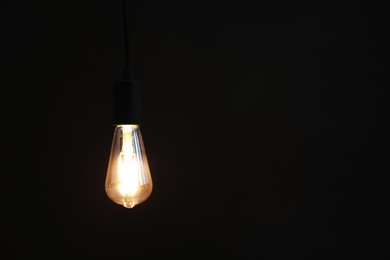 Photo of Glowing light bulb hanging on dark background. Space for text