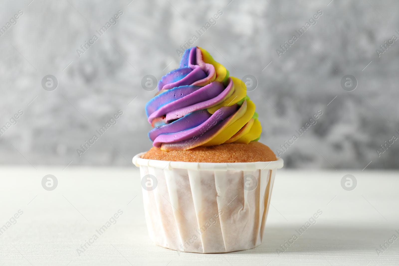 Photo of One delicious cupcake with colorful cream on white wooden table against blurred grey background, closeup