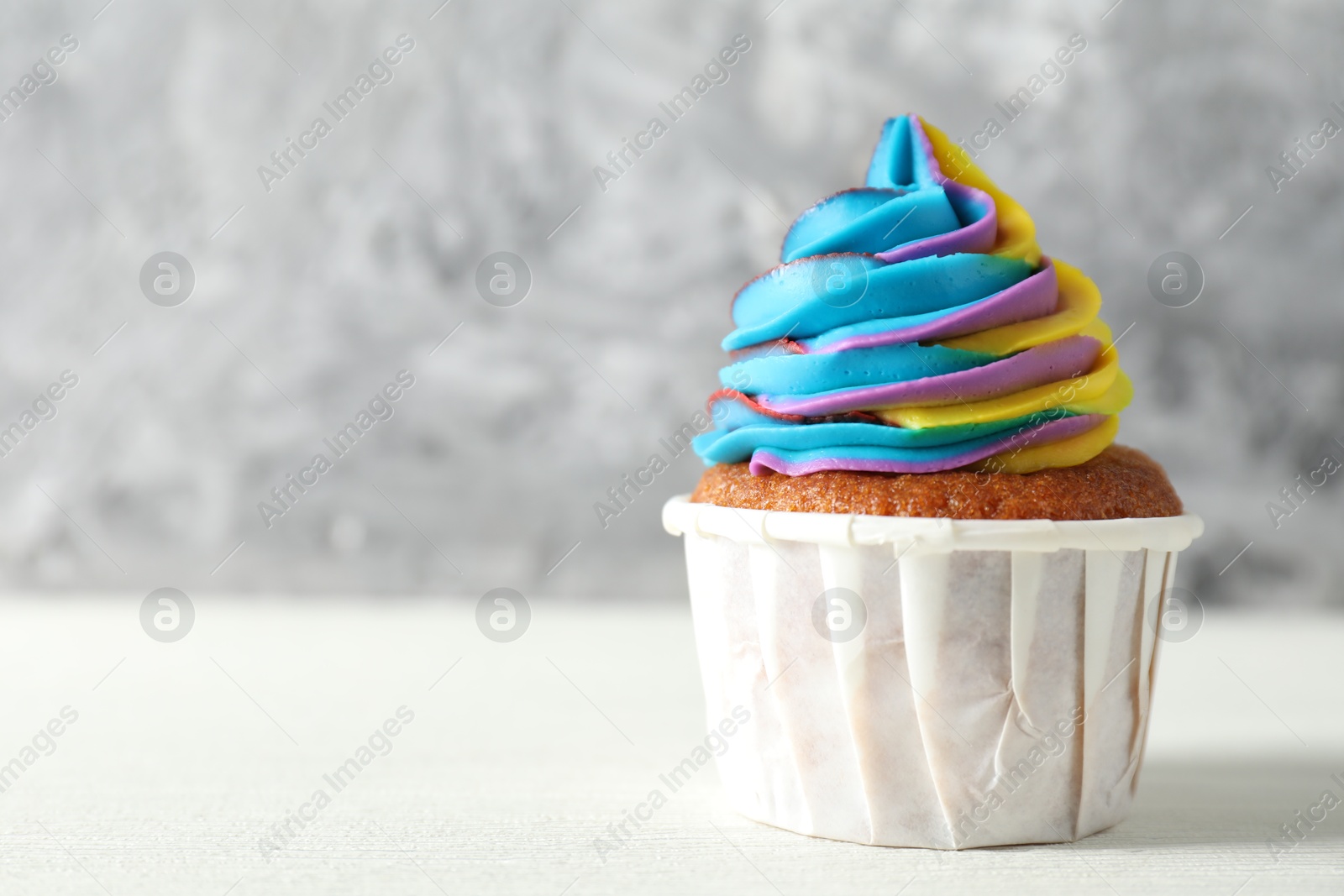 Photo of One delicious cupcake with colorful cream on white wooden table against blurred grey background, closeup. Space for text