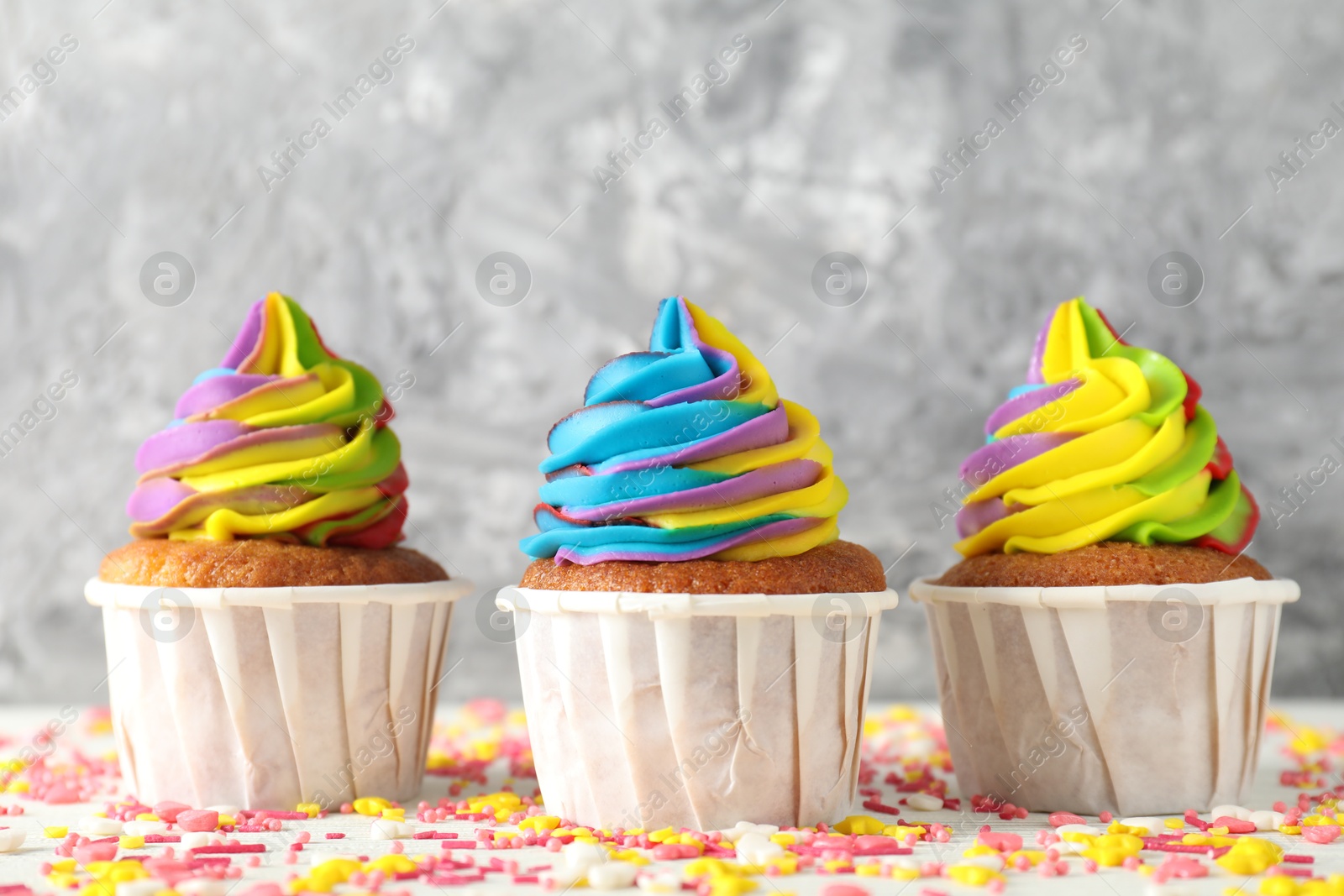 Photo of Delicious cupcakes with colorful cream and sprinkles on white table against blurred grey background, closeup