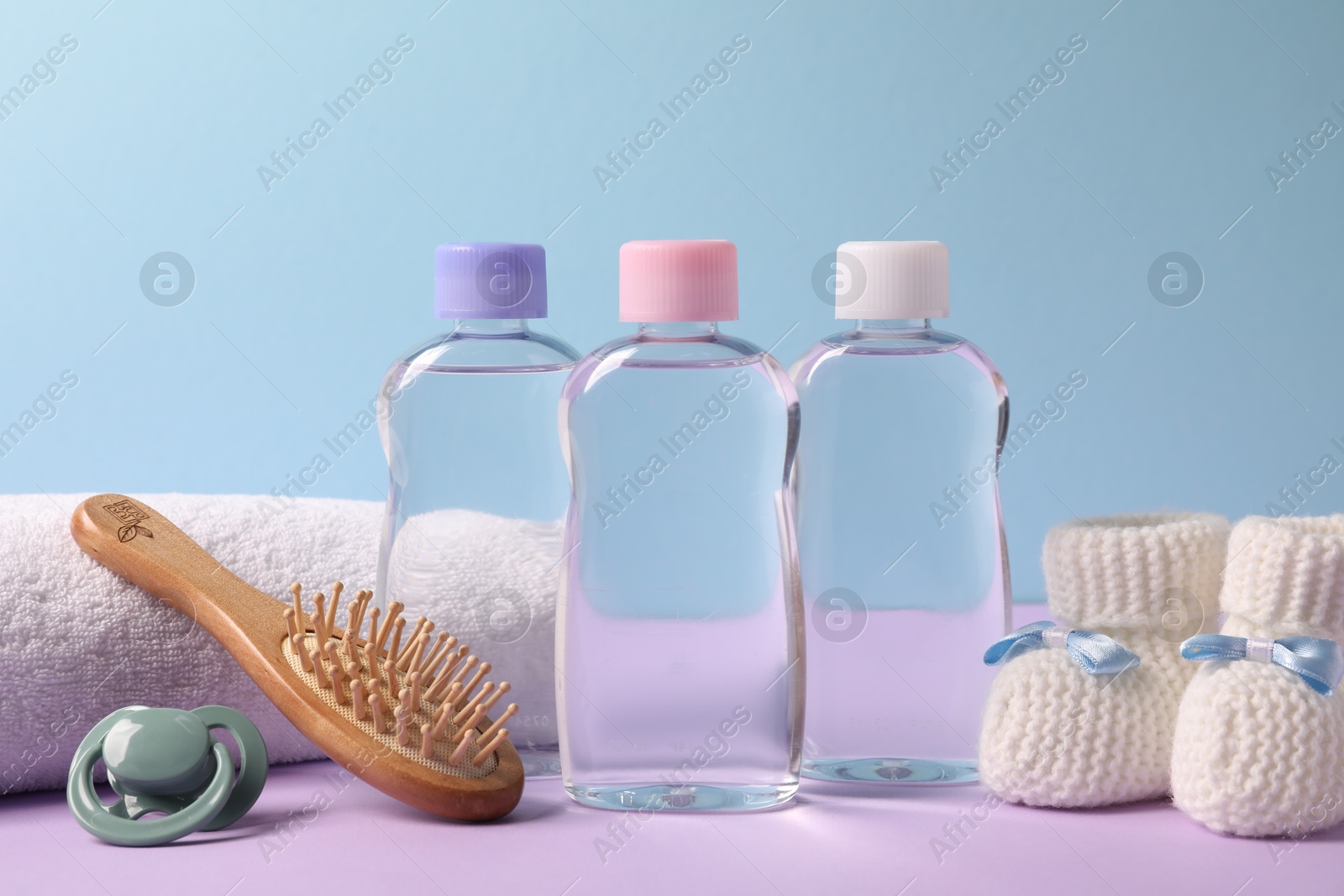 Photo of Baby oil in bottles, pacifier, brush and accessories on violet table against light blue background, closeup