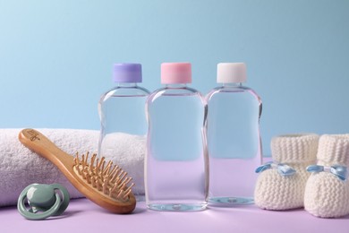 Photo of Baby oil in bottles, pacifier, brush and accessories on violet table against light blue background, closeup