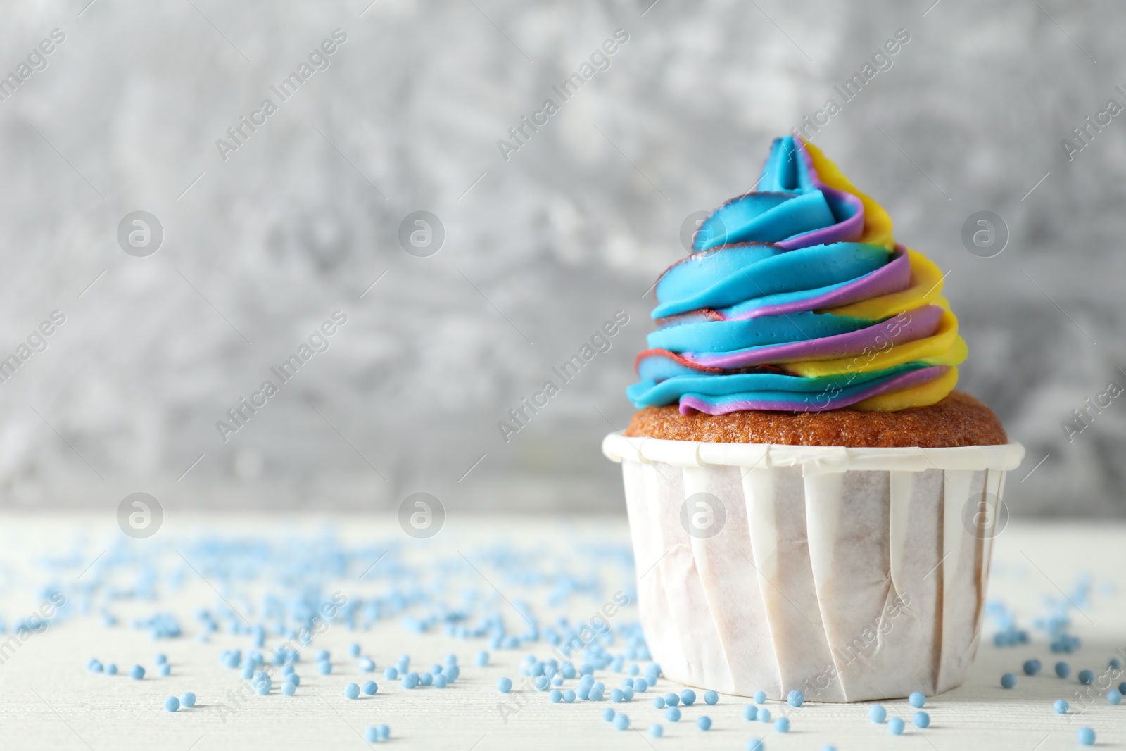 Photo of One delicious cupcake with colorful cream and sprinkles on white wooden table against blurred grey background, closeup. Space for text