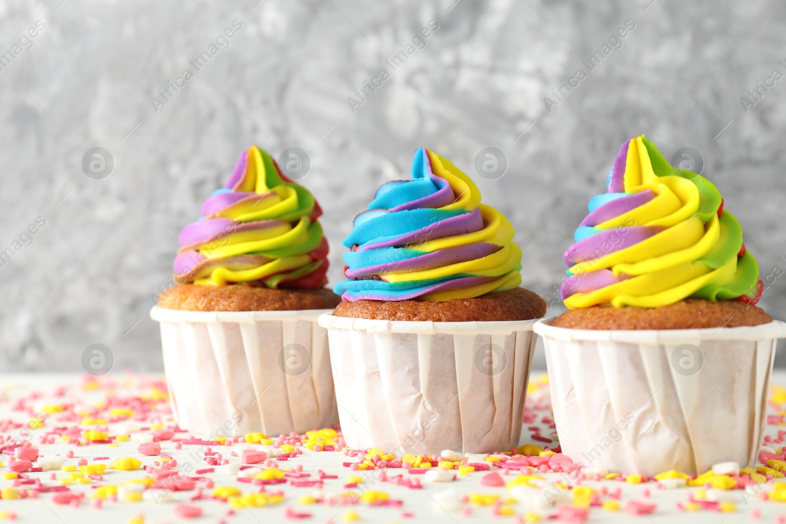 Photo of Delicious cupcakes with colorful cream and sprinkles on white table against blurred grey background, closeup