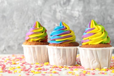 Photo of Delicious cupcakes with colorful cream and sprinkles on white table against blurred grey background, closeup